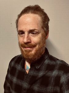 A white male with red hair and red beard, brown eyes, and wearing a black and white collared shirt.  