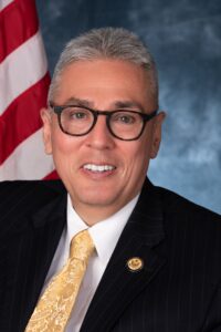 Caucasian male with black rimmed eyeglasses, short greying hair, a dark suit jacket with a white dress shirt and yellow tie.