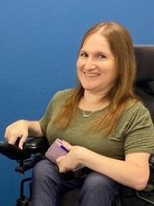 A person with long hair using a power wheelchair smiles toward the camera