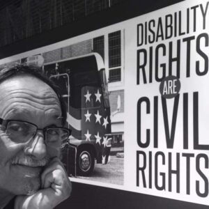 A man with his hand on his face, wearing glasses and smiling for the camera in front of a banner with a bus and the words Disability Rights Are Civil Rights