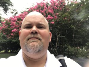 A white middle aged male with a close cropped hair and goatee wearing a white polo shirt, standing in front of what looks like pink cherry blossoms.