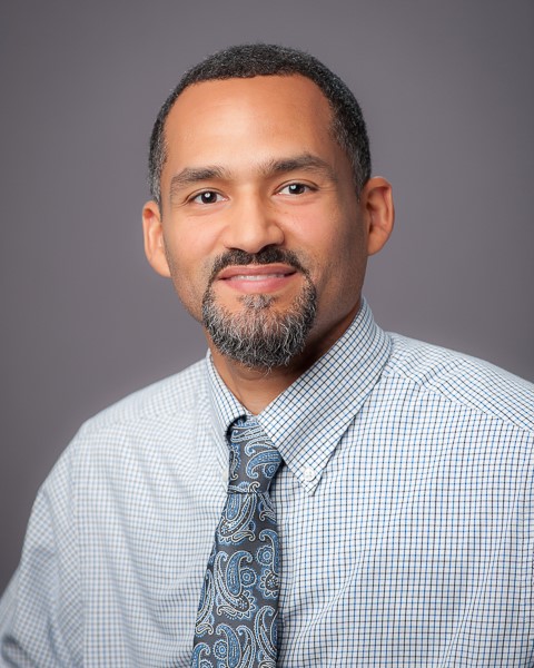 Professional headshot- Latino male, short dark hair and goatee, wearing a plaid button-down shirt with a paisley tie.