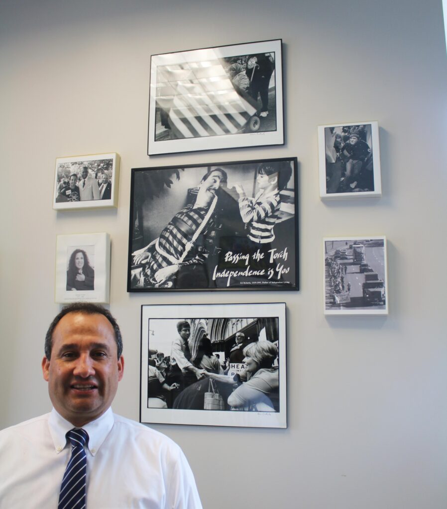 Liberty Resources Inc. CEO Thomas H. Earle smiles in front of a wall of black and white disability rights activism photos that span decades. Mr. Earle is a black-haired Latino man with a white Oxford shirt and black and white striped tie. 