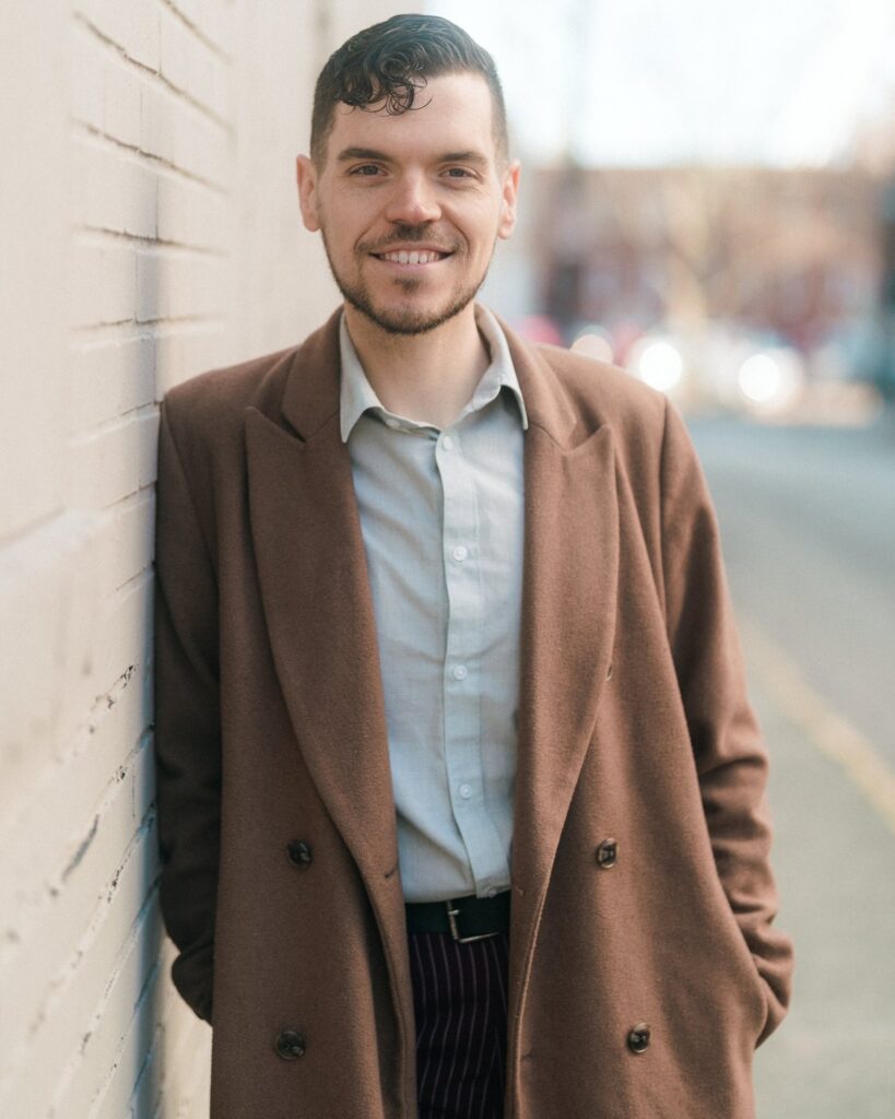 A photo of a white man in his 30s with dark brown hair wearing a light brown coat smiling and leaning against a white wall