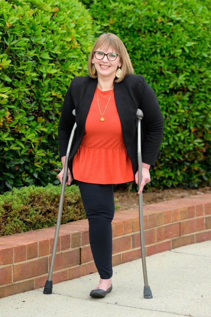 Caucasian, blond woman with glasses, black jacket/pants and red top. She is smiling, has one leg and is using crutches. 