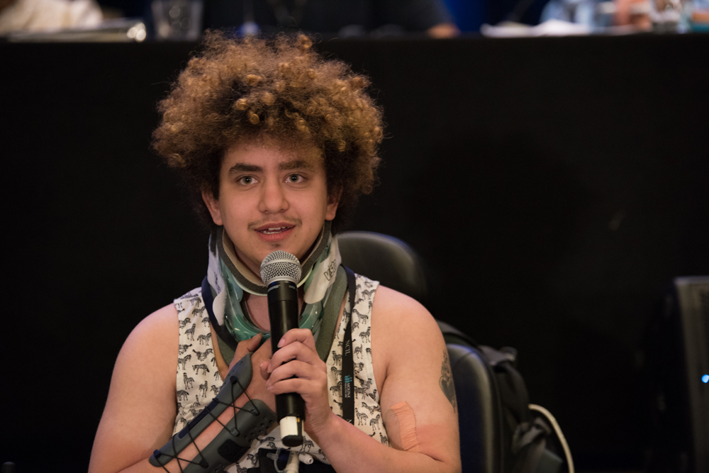 Emet Tauber speaks into a microphone during NCIL’s Annual Conference. Emet has curly hair and is wearing a tank top and neck brace. 
